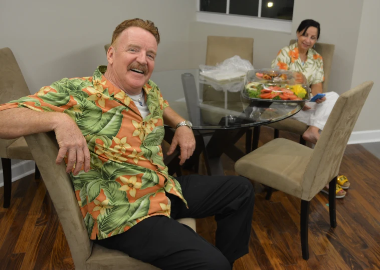 a man in hawaiian shirt sitting at a table with woman behind him