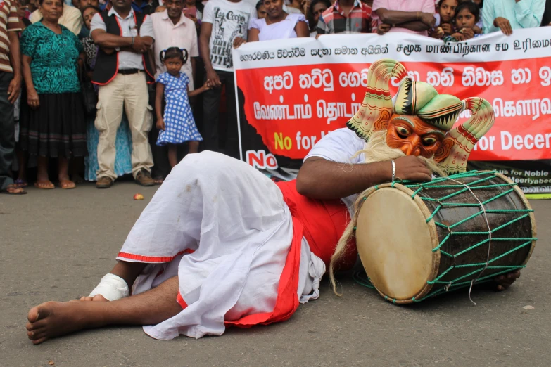 an old man laying on the ground in front of a crowd with a mask and musical instrument