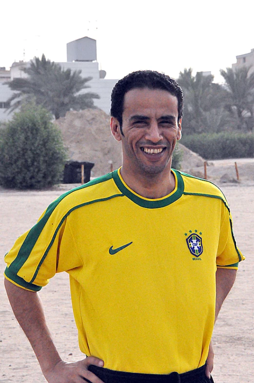 a smiling man stands on the beach wearing soccer shirt