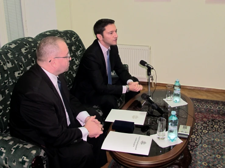 two men in suits sitting on a chair while holding papers and a microphone