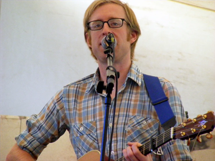 a man with glasses and guitar sings into a microphone