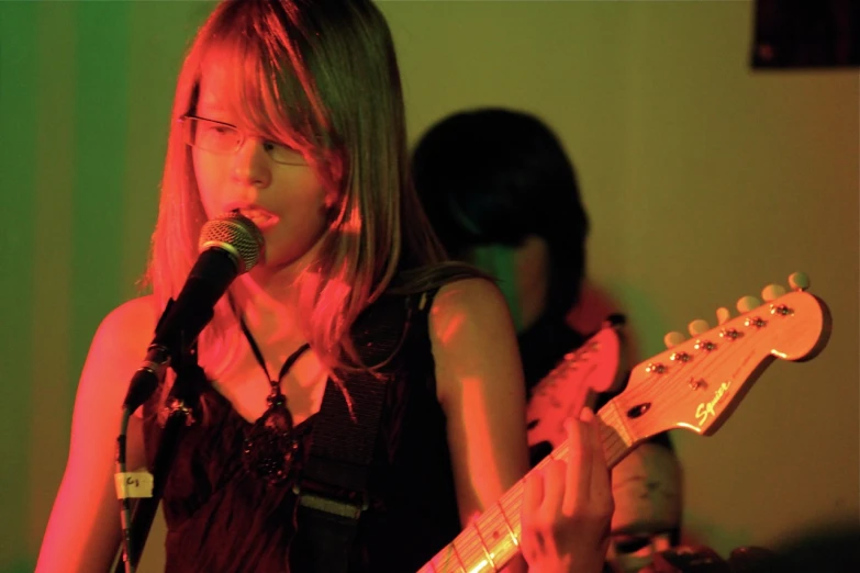 a girl with glasses playing a guitar while another person is singing behind her