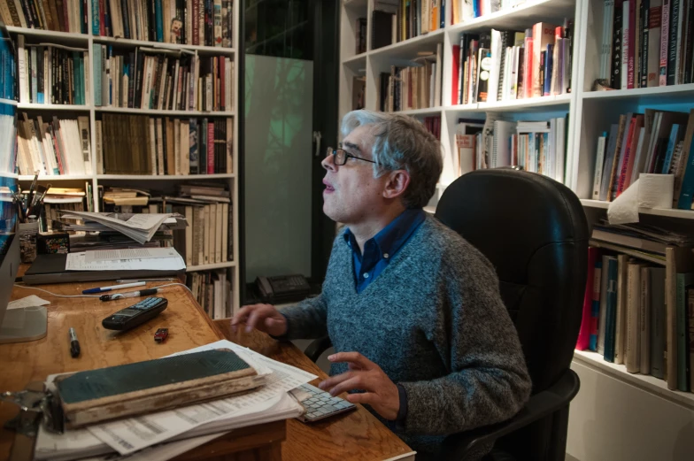 an older man sitting at a desk in front of a bookcase with books