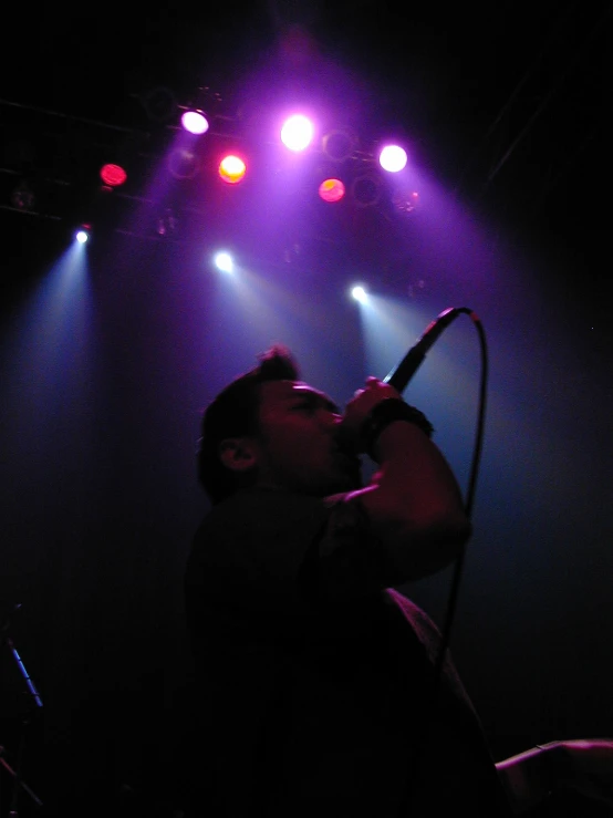 man on stage singing with a microphone in front of purple and red lights