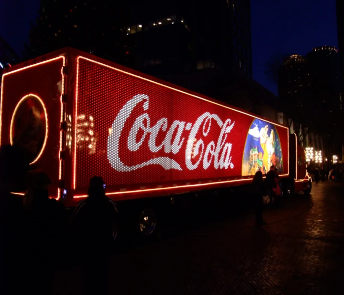 an illuminated coca - cola truck is parked on the street at night