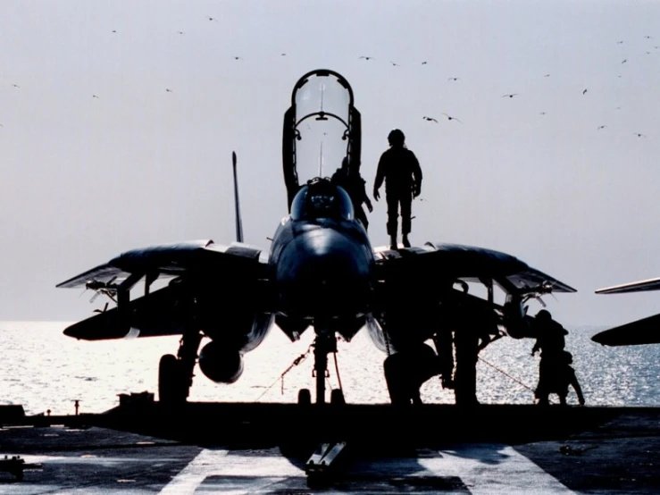 a navy plane sits on the flight deck while men stand in front of it