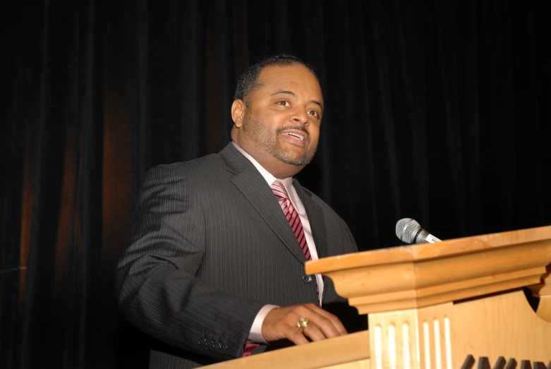 a man wearing a suit and tie is speaking at a podium