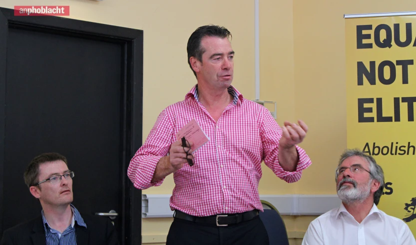 man in pink shirt speaking at an event with men sitting around