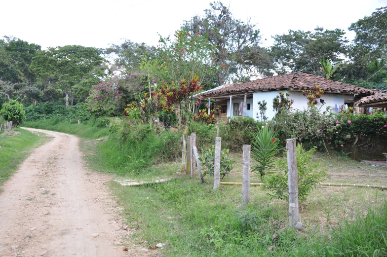 the dirt road has an old building on one side and a stone roof and a broken fence around it