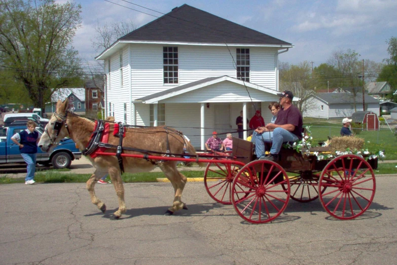 two people on a buggy pulling a horse drawn carriage with many people standing outside