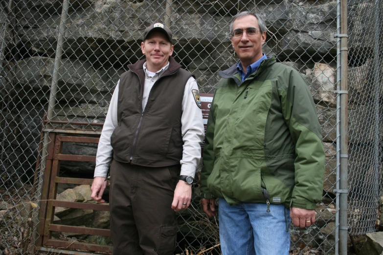 two men standing in front of a stone wall with a fence behind them