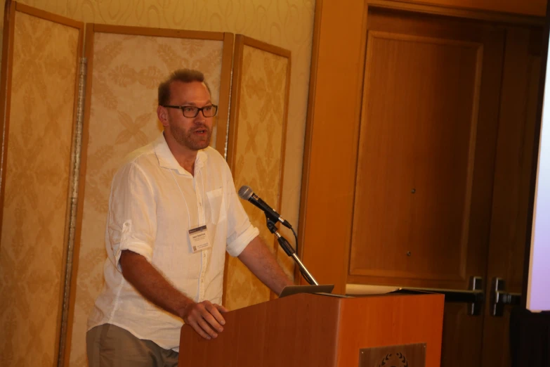 a man in glasses stands at a podium in front of a projection screen