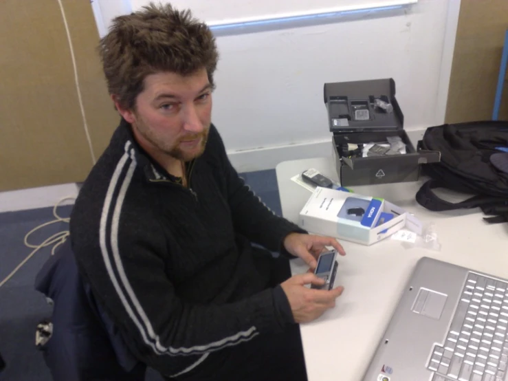 a man with beard and a jacket on sitting at a desk in front of a laptop