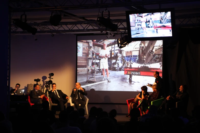 a crowd of people are sitting on a stage in front of a projection screen