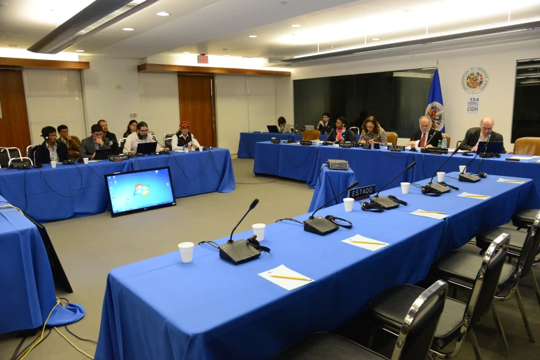 a large table set up with microphones and people sitting at a long blue desk