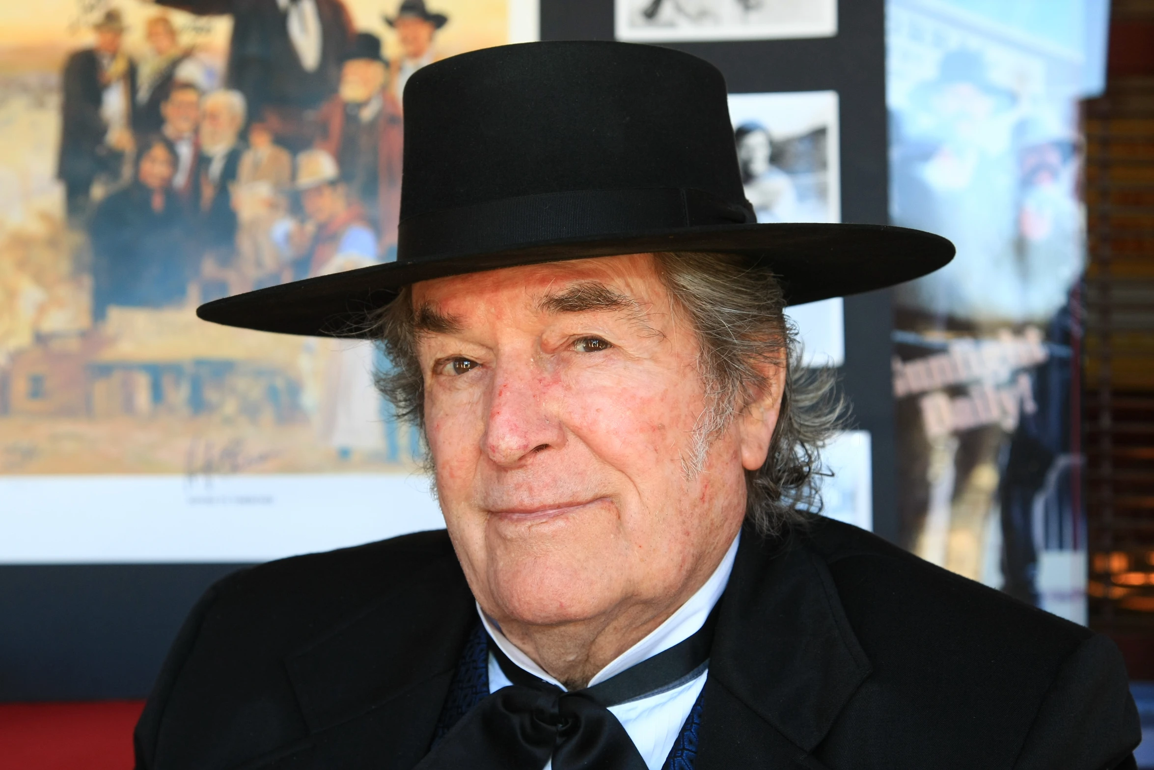 man with grey hair wearing a black suit and fedora