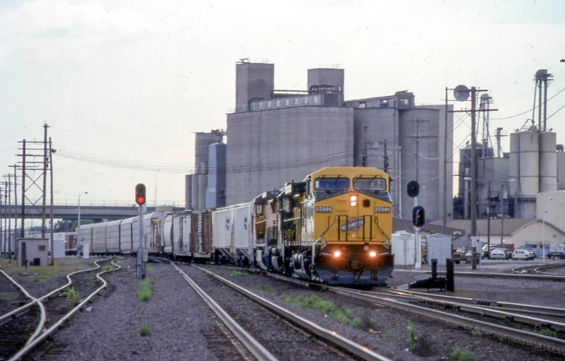 yellow train with several cars on tracks in front of factory building