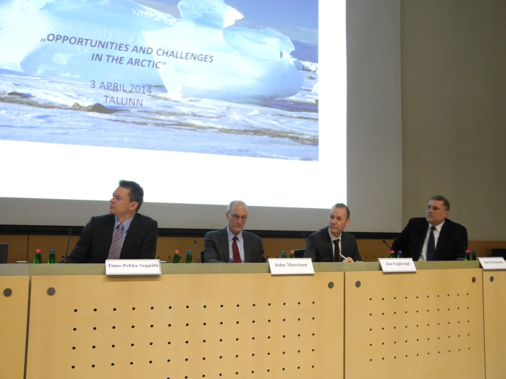 five men sitting at tables in front of a projection screen