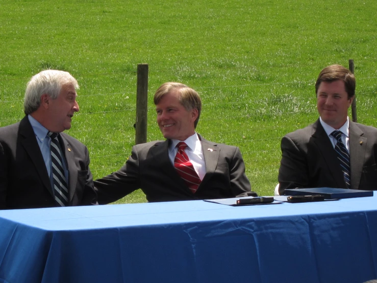 three men sit at a table near some grass