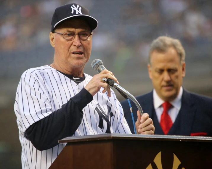 a man wearing glasses speaking at a podium while wearing a baseball jersey