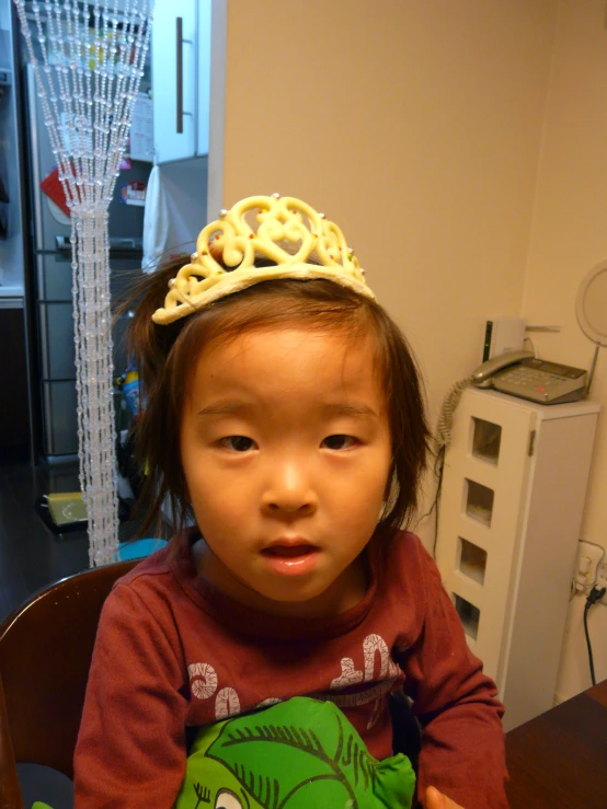 an asian girl in a gold crown holds a green frog pillow