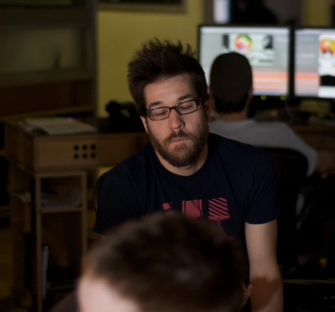 a bearded man wearing glasses sitting in front of three computer screens