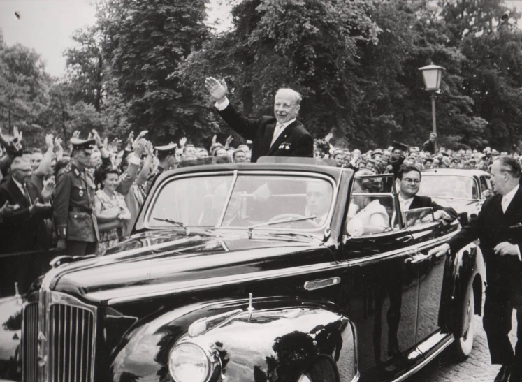 a man waving from an old black car and a crowd is watching