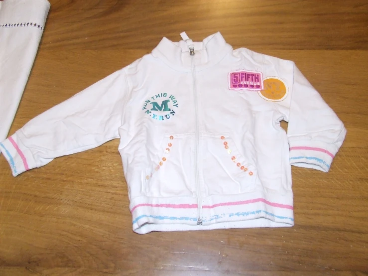 a child's white jacket with a patch in the front and a hat on the back