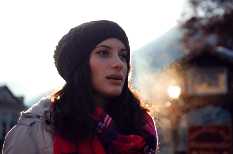 a young woman in winter clothes stands against a background of town houses and trees