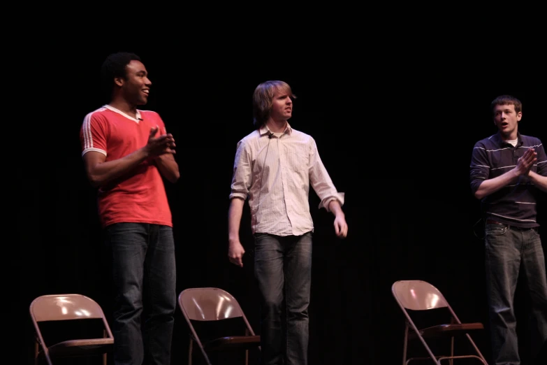 three young men standing on stage one of them wearing an open collar shirt