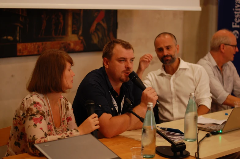 three people sit at a table with water bottles, a laptop, a microphone and two other people are around