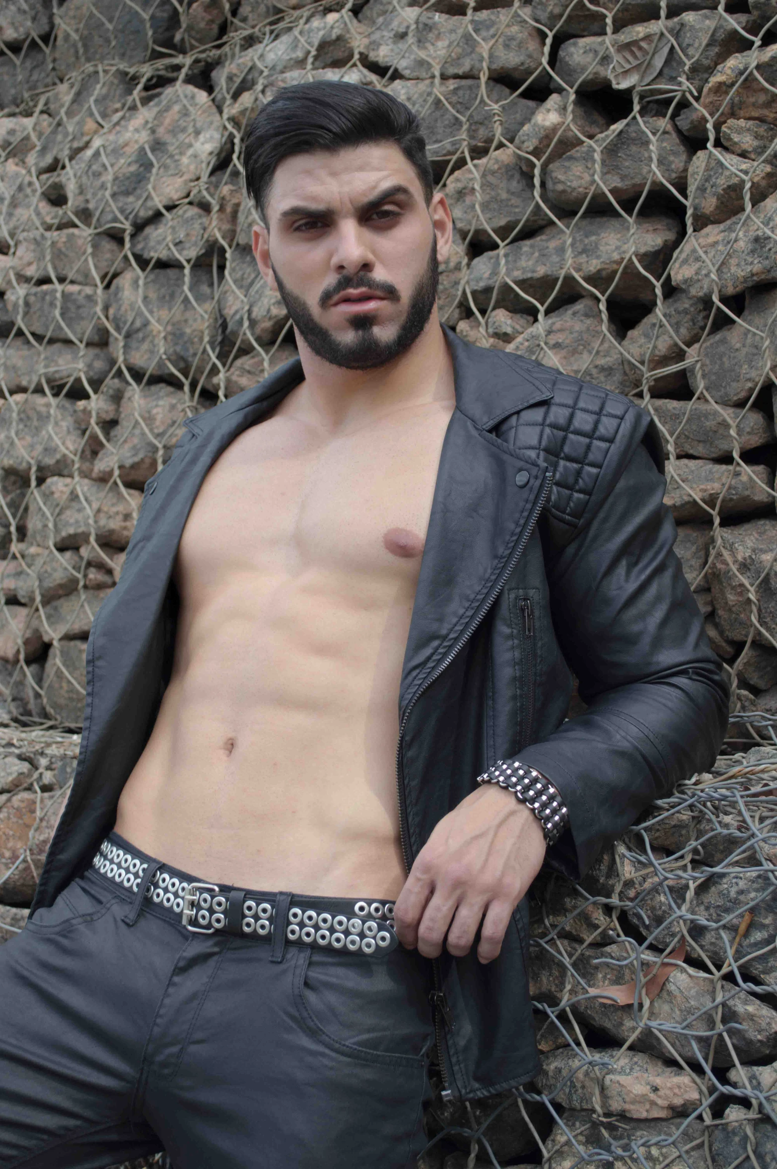 shirtless man in leather and black jacket standing with a chain link fence behind him