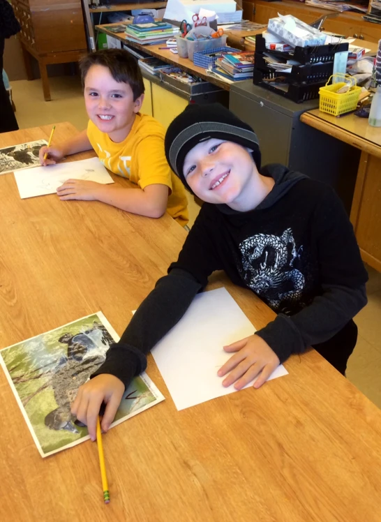 two boys smiling while sitting at a wooden table