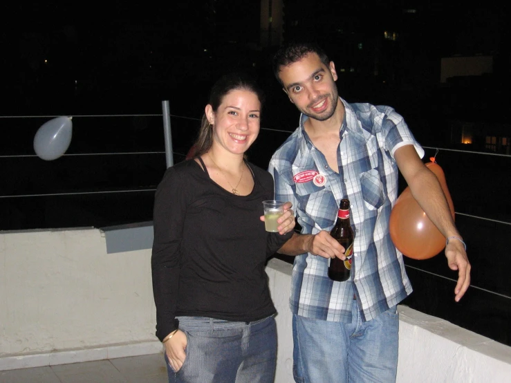 a couple holds a glass and a drink as they stand on a deck at night