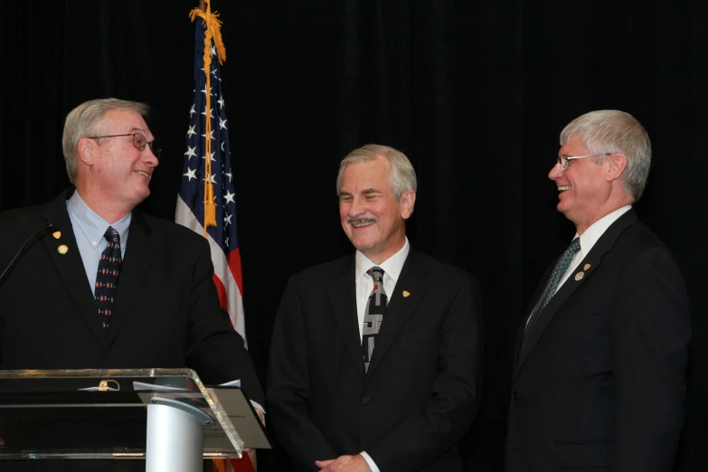 three men in suits stand at a podium