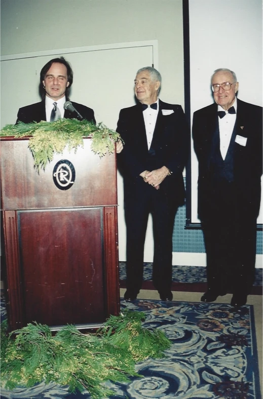 two men in suits and ties standing near a podium with a plant at the front