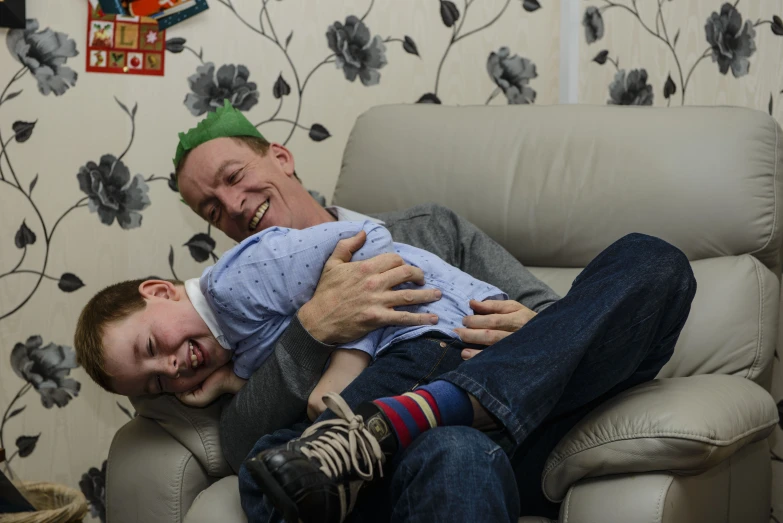 a man sitting on a couch being hugged by a man in front of flowers on a wall