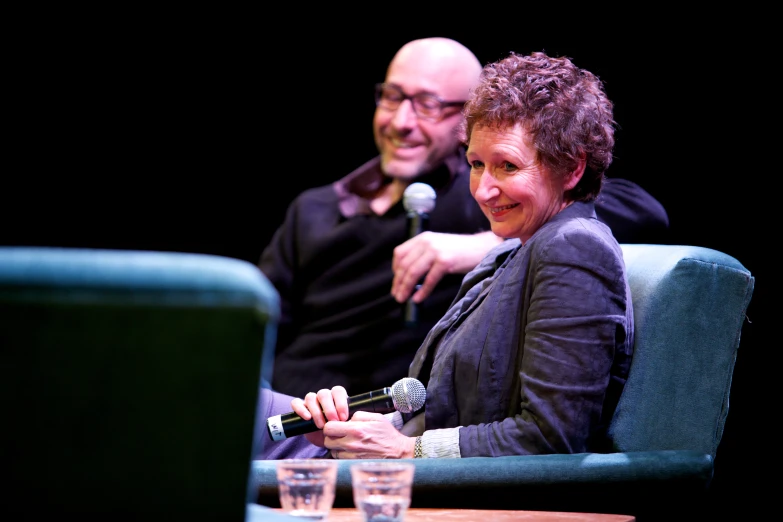two people are sitting in a stage or audience, one of which is holding a microphone, the other a laptop