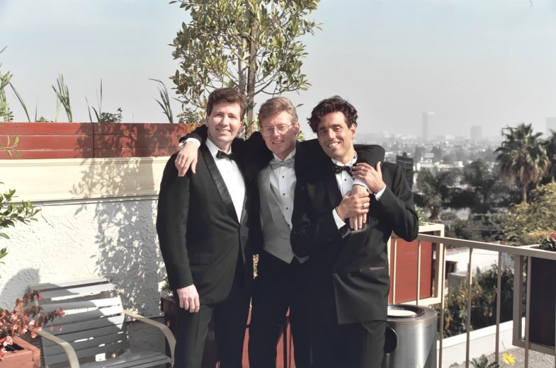 three young men in tuxedos and bow ties posing together on a rooftop with city skyline behind them