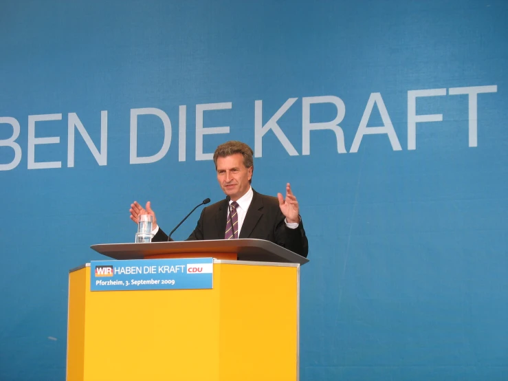 man giving speech at podium with sign in background