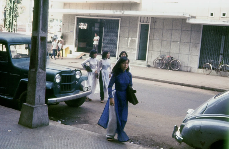 people walk down the sidewalk near an old car