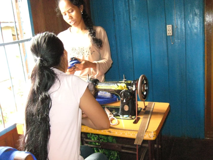 a girl sitting next to another woman working on a sewing machine