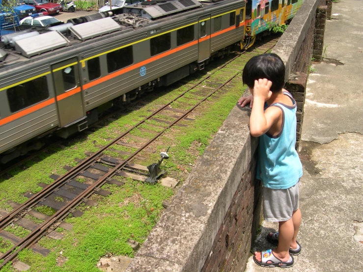 a little boy leaning against the rail wall with his hand to his head as he looks at train cars and cars