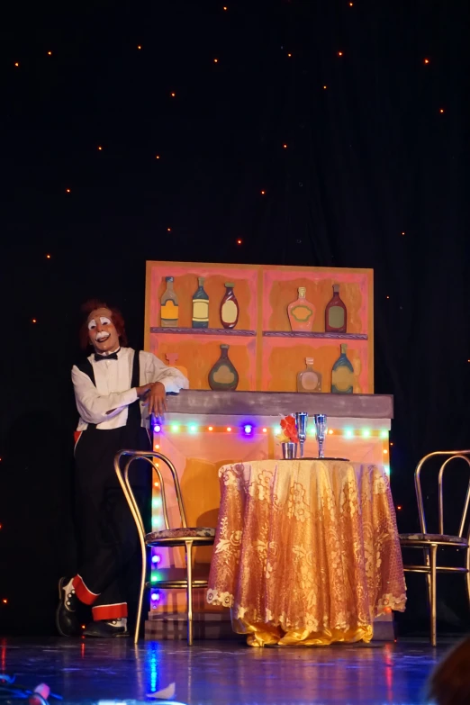 a masked man sits at a table in front of a podium with wine bottles on it