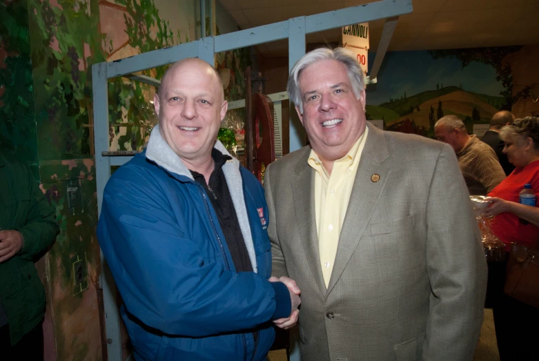 two older men smile at the camera while one shakes hands with another