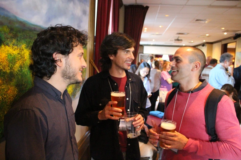two men laugh as they hold beers in front of a group of people