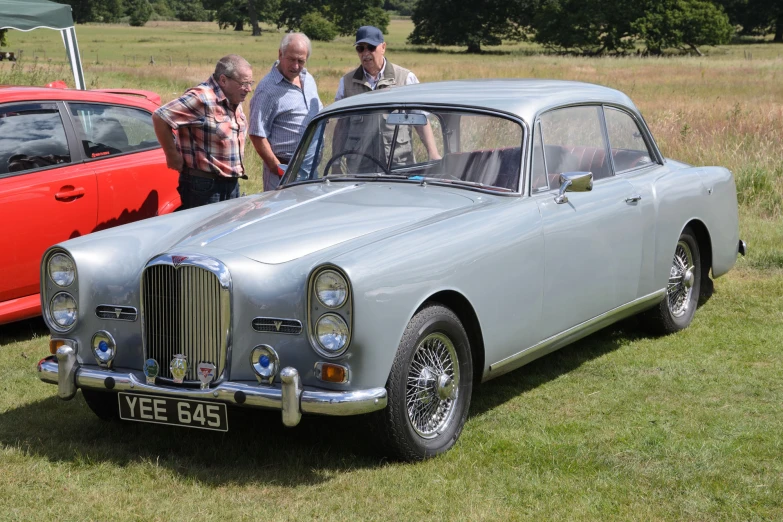 the two men stand next to two classic cars