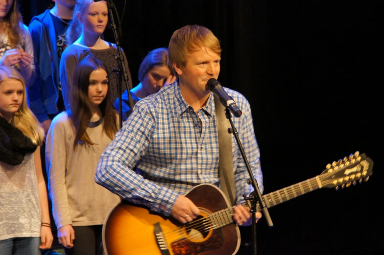 a man sings into a microphone while standing on stage with his guitar