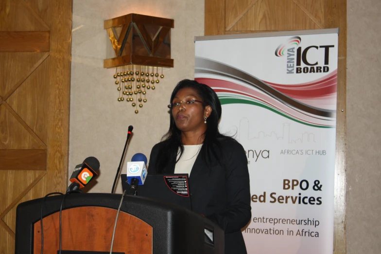 a woman in black jacket at podium holding microphone in front of two speakers