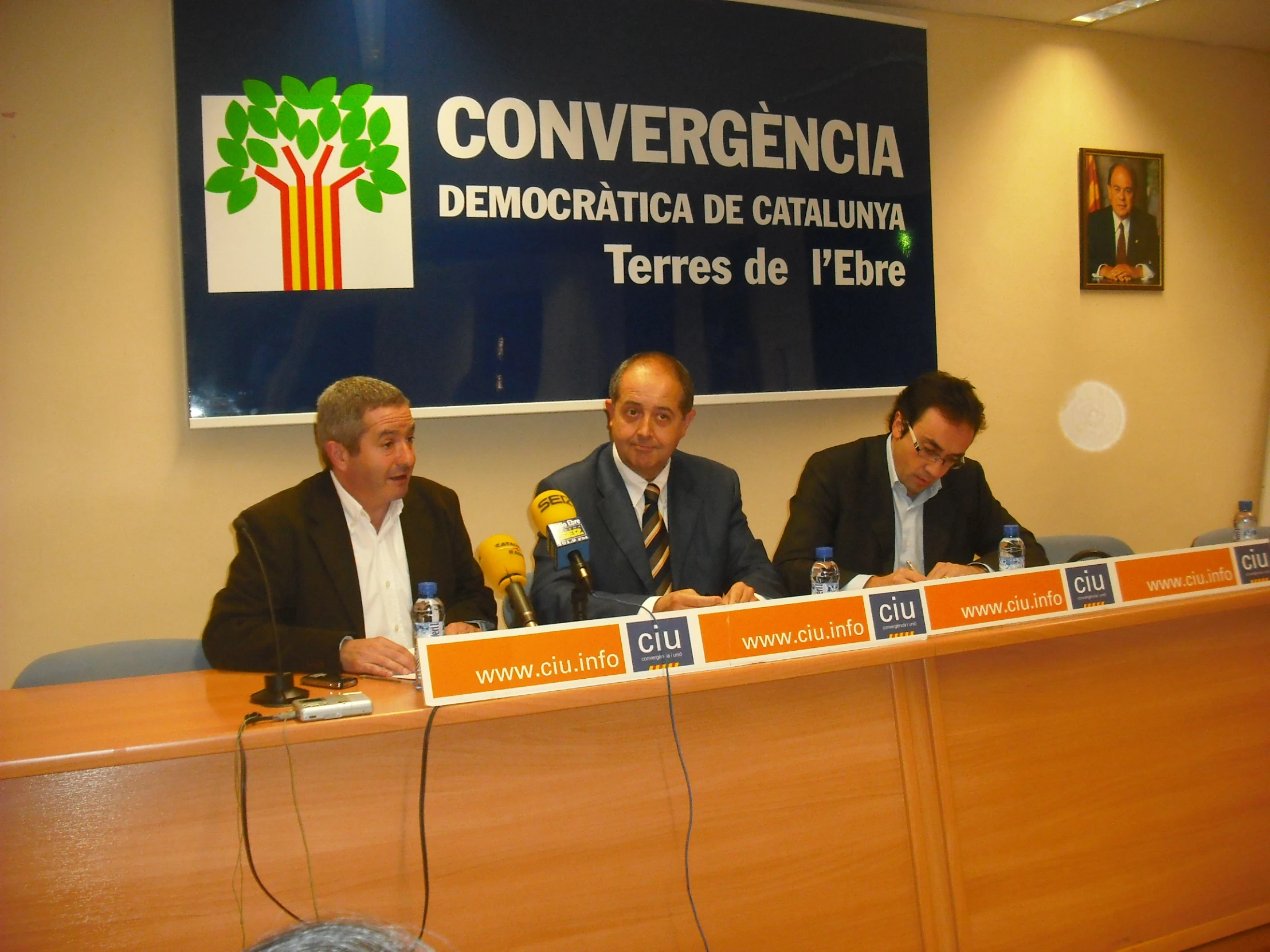 three men sitting at a table speaking in front of a sign